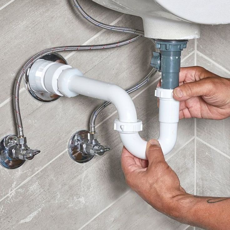 Mãos instalando sifão e conexões de esgoto sob uma pia, com registros e flexíveis de água